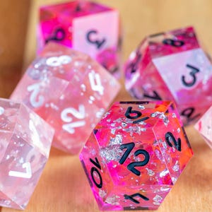 Pode incluir: Vários dados poliédricos com um design de resina rosa e transparente. Os dados têm números pretos e detalhes em folha de prata. Estão sobre uma superfície de madeira castanha clara. Para jogos de tabuleiro.