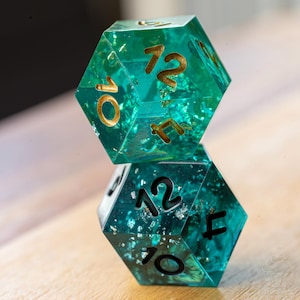 May include: Two hexagonal dice, one atop the other, with a teal resin base and gold-colored numbering. The dice have a glittery, speckled appearance. The numbers 10, 12, and 4 are visible on the dice.