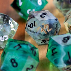 May include: Close-up of several polyhedral dice with a marbled effect. The dice are primarily green and clear with black and gold numbering. The dice are used for tabletop role-playing games.