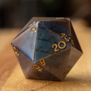 May include: A multi-faceted die with gold numerals and text. The die has a dark, marbled appearance with blue and brown tones. The text on the die reads "true happiness, loved, & what".
