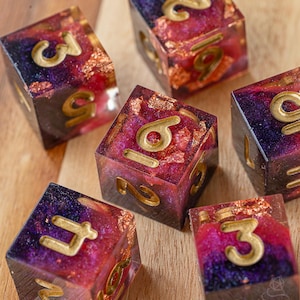 Pode incluir: Seis dados artesanais com tema celestial. Cada dado é um cubo com números dourados e uma mistura de roxo, vermelho e folha de ouro embutidos em resina transparente. Os dados estão dispostos sobre uma superfície de madeira.