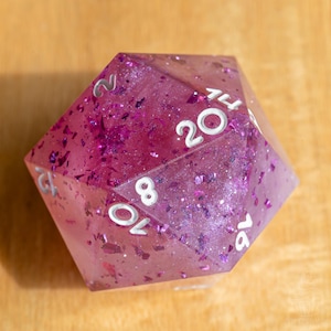 May include: A translucent twenty-sided die in a vibrant magenta hue, featuring embedded purple metallic flakes and shimmering glitter throughout the resin. The numbers are clearly engraved and filled with a crisp white ink for high contrast.