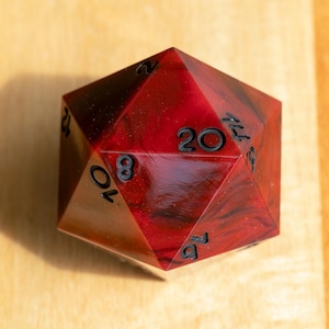 May include: A twenty-sided die featuring a marbled red and black resin finish with subtle glitter accents. The die has sharp geometric edges and clear, recessed black numerals engraved into the faces, including the number 20 at the top centre.