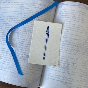 May include: A pen illustration on a small card, with a blue ribbon bookmark and an open book. The pen is blue and white, and the card has a textured surface. The book pages are visible with text.