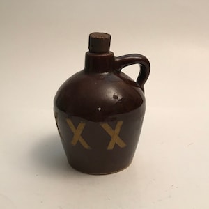 May include: A brown ceramic jug with a cork stopper and a handle. The jug has the letters "XXX" written on the front in a yellow color.