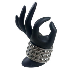 May include: A black hand-shaped jewelry display holding a silver bracelet. The bracelet features square, gray-toned studs and a row of smaller, clear stones. The display is set against a white background.
