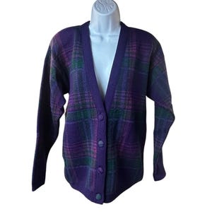 May include: A long-sleeve V-neck cardigan sweater featuring a purple, green, and dark blue plaid pattern. The garment has a solid purple button-front placket with four matching circular buttons, long sleeves, and a ribbed hem and cuff detail.