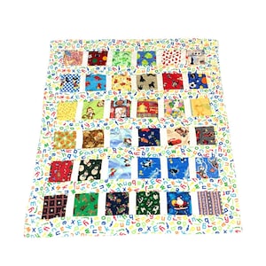 May include: A rectangular quilt featuring a white background printed with a colorful alphabet pattern. The quilt is composed of six rows of six square fabric patches, each displaying various cartoon characters, holiday themes, and geometric prints.