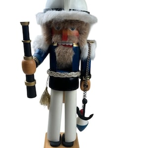 May include: A wooden nutcracker figure dressed as a sailor, wearing a white helmet with white faux fur trim, a blue jacket, and white trousers. The figure holds a black telescope in one hand and has a small black anchor charm hanging from a chain.