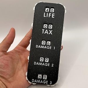 Könnte beinhalten: Ein schwarzer Spielzähler mit weißem Text und Zahlen. Der Zähler hat die Wörter "LIFE", "TAX" und "DAMAGE" mit numerischen Anzeigen. Die Kanten sind mit einem weißen, gewellten Design verziert. Der Zähler wird in einer Hand gehalten.