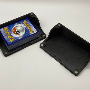 Peut inclure: Une boîte de rangement en plastique noir avec un couvercle à charnière, ouverte pour révéler une pile de cartes à jouer Pokémon. La boîte a une finition texturée et est conçue pour contenir un jeu de cartes standard.