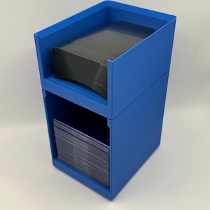 May include: A two-tiered, blue storage container, likely for cards or small items. The top section is angled, and both sections hold stacks of dark-colored cards. The container is made of a solid, opaque material.