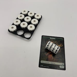 May include: A black plastic tray holding ten white circular counters with black numbers, alongside a rectangular game card labelled Squirrel with a 1/1 token creature designation and three stacked white numbered counters.