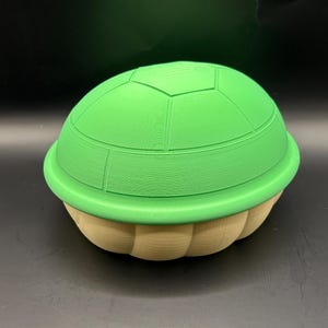 May include: A green turtle shell with a light tan base. The top of the shell is a bright green with a segmented pattern. The base is a light tan color with a ribbed texture. The shell is a 3D-printed item.