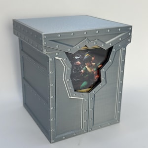 May include: A gray, riveted box with a hinged lid. The box has a decorative cutout revealing a colorful image of a character. The box is designed to hold cards or small items.
