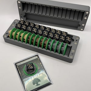 May include: A grey plastic storage case containing rows of black dice with gold +1/+1 markings and green and brown circular tokens. A single black die with gold +1/+1 markings sits on a green token atop a Forest card in a clear plastic sleeve.
