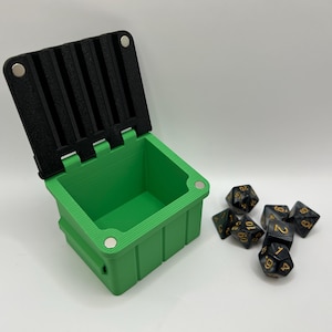 Op de afbeelding: Een groene, magnetische dobbelstenendoos met een zwart deksel. Het deksel is open en er liggen verschillende zwarte dobbelstenen met gouden cijfers op een wit oppervlak.