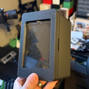 以下が含まれることがあります： 透明な窓が付いた黒い長方形のディスプレイケース。ケースはテクスチャ加工された表面と側面の小さなスロットを備えています。ケースは人の手に持たれています。