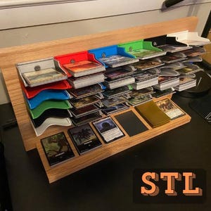 May include: Wooden card organizer with multiple shelves in various colors, including red, blue, green, and black. The organizer holds stacks of trading cards, with some cards displayed on the bottom shelf. The letters "STL" are visible in the bottom right corner.