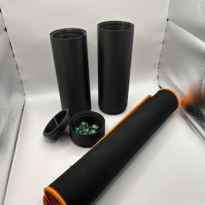 以下が含まれることがあります： ふた付きの黒い円筒形の容器が2つ、1つは緑色のサイコロが入っており、もう1つは黒とオレンジのロールアップされたゲームマットです。