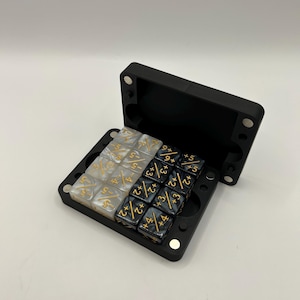 May include: A black, hinged dice case with a magnetic closure, open to reveal two sets of dice. One set is pearlescent with gold markings, the other is black with gold markings. The dice are for tabletop games.