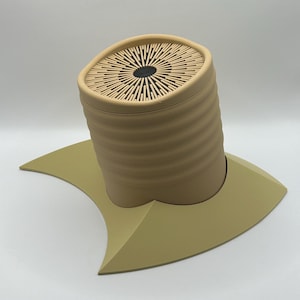 May include: A beige ceramic container with a textured, ribbed design and a circular lid with a black center. The container is sitting on a beige, crescent-shaped base.
