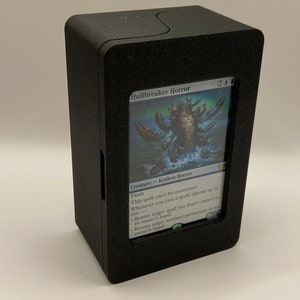 Può includere: Una scatola di gioco di carte nera con una finestra trasparente che mostra una carta Magic: The Gathering blu e bianca con il testo "Hullbreaker Horror" e un'immagine di una grande creatura blu simile a un granchio. Il testo della carta dice "Creature - Kraken Horror".