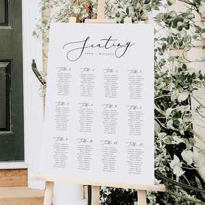 May include: A white wedding seating chart with black calligraphy lettering. The chart lists guest names by table number. The chart is titled "Seating" with the names "Sara + Michael" below the title.
