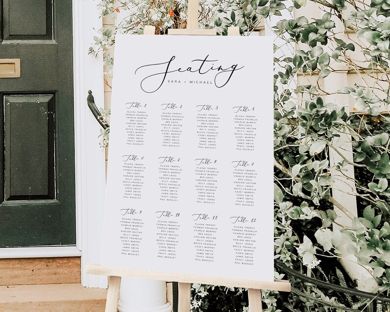 May include: A white wedding seating chart with black calligraphy lettering. The chart lists guest names by table number. The chart is titled "Seating" with the names "Sara + Michael" below the title.