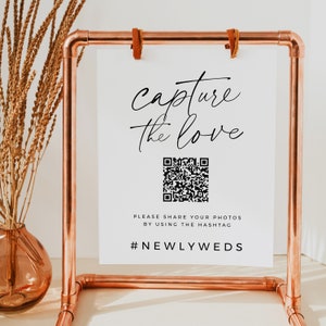 Könnte beinhalten: Ein weißes Schild mit dem Text "capture the love" in schwarzer Kursivschrift. Unter dem Text befindet sich ein schwarz-weißer QR-Code. Das Schild enthält auch den Text "Please share your photos by using the hashtag #NEWLYWEDS". Das Schild wird auf einem Ständer aus Kupferrohren präsentiert.