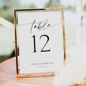 May include: Gold-framed table number sign with the text "table 12" in black script font. Below the number is the text "LAURA & MARTIN 11.09.XX" in black.