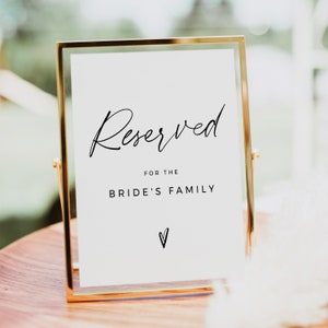 May include: A framed sign with the word "Reserved" in elegant script, followed by "for the Bride's Family" in a simple font. A small heart symbol is at the bottom. The frame is gold, and the sign is on a wooden surface.