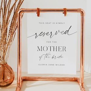 May include: A white sign with black text on a copper pipe stand. The sign reads "This seat is kindly reserved for the Mother of the bride Gloria Jane Wilder".