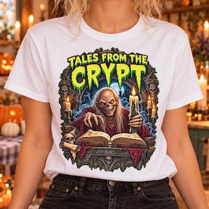 May include: A white t-shirt featuring a graphic print of the Crypt Keeper from Tales from the Crypt. The design shows a skeletal figure in a red robe reading an open book by candlelight, surrounded by stone arches, skulls, and green text.