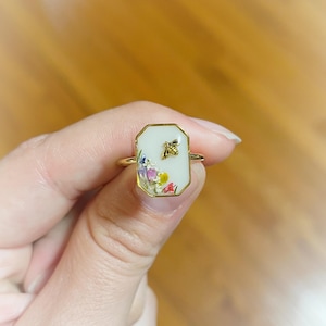 May include: A gold-toned ring with a rectangular, off-white face. The face features a small, gold-colored bee and a collection of colorful, dried flowers. The ring is held up against a blurred wooden background.