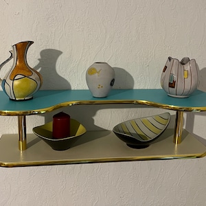 Peut inclure: Une étagère à deux niveaux avec un dessus turquoise et un dessous beige, tous deux bordés d'or. L'étagère supérieure contient une cruche décorative, un petit vase et un vase en forme de fleur. L'étagère inférieure présente une bougie et deux bols décoratifs.