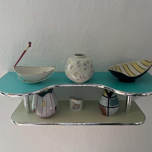 May include: A two-tiered wall shelf with a turquoise top shelf and a light gray bottom shelf, both featuring chrome edges and supports. The top shelf holds a white bowl with a red accent, a round vase with pastel patterns, and a yellow striped bowl.
