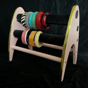 Op de afbeelding: Een roze houten sieradenstandaard met gouden rand, met een verscheidenheid aan kleurrijke armbanden. De standaard heeft een modern design uit het midden van de eeuw met twee zwarte fluwelen staven om de armbanden vast te houden.