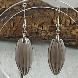 May include: A pair of gray, ribbed oval earrings with silver-colored hooks. The earrings are displayed on a clear stand against a blurred wooden background. The earrings have a vertical striped design.