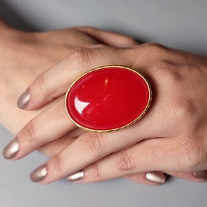 May include: A large, oval-shaped ring with a bright red stone set in a gold-toned metal frame.