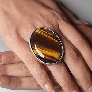 May include: A silver ring featuring a large oval tiger's eye gemstone. The gemstone is brown and gold in colour with a shimmering effect.