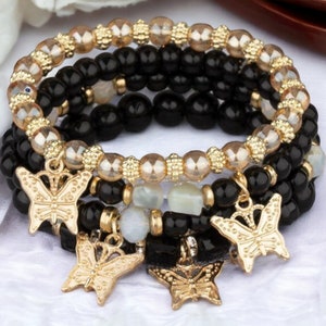May include: A set of beaded bracelets in black and gold. The bracelets have gold butterfly charms. The beads are round and smooth. The bracelets are stacked, creating a layered effect. The image shows the bracelets on a white surface.