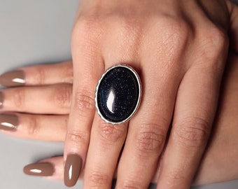 May include: A silver ring with a large oval-shaped blue stone set in a silver bezel. The stone has a sparkly, glittery texture.