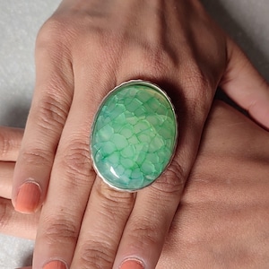 May include: A large oval ring with a silver band. The ring features a light green stone with a textured, cracked pattern. The stone is set in a silver bezel. The ring is on a hand with orange nail polish.