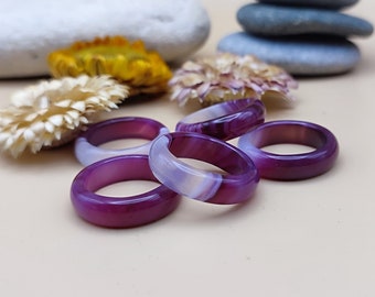 May include: Five purple and white agate rings stacked on a light beige surface. The rings are made of natural stone and have a polished finish.