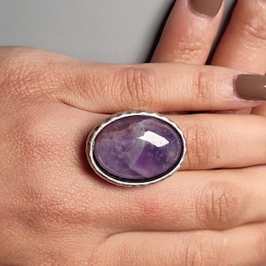 Könnte beinhalten: Ein silberner Ring mit einem großen ovalen Amethyst-Edelstein. Der Ring befindet sich an einem Finger.