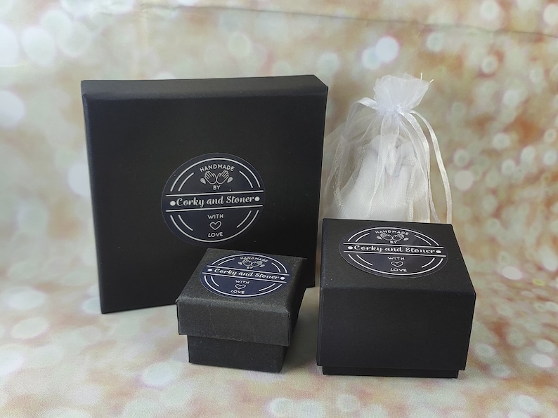 Pode incluir: Tr&ecirc;s caixas pretas com tampas, cada uma com um adesivo circular branco que diz "Handmade by Corky and Stoner. With love." As caixas est&atilde;o sobre uma superf&iacute;cie marrom claro.