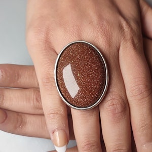 May include: A silver ring featuring a large oval brown stone set in a silver bezel. The stone has a sparkly, glittery texture.