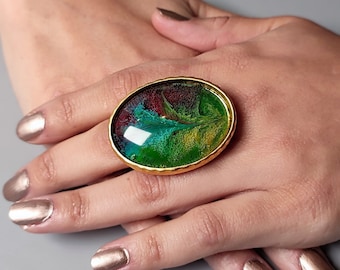 May include: An oval ring featuring a gold-toned metal setting with a textured edge. The centre is filled with a glossy, resin-like material displaying a vibrant, swirling pattern of deep green, bright lime, teal, and dark burgundy colours.