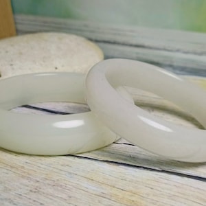May include: Two white jade bangles, one slightly larger than the other, are displayed on a light wood surface. The bangles are smooth and polished, with a subtle sheen.